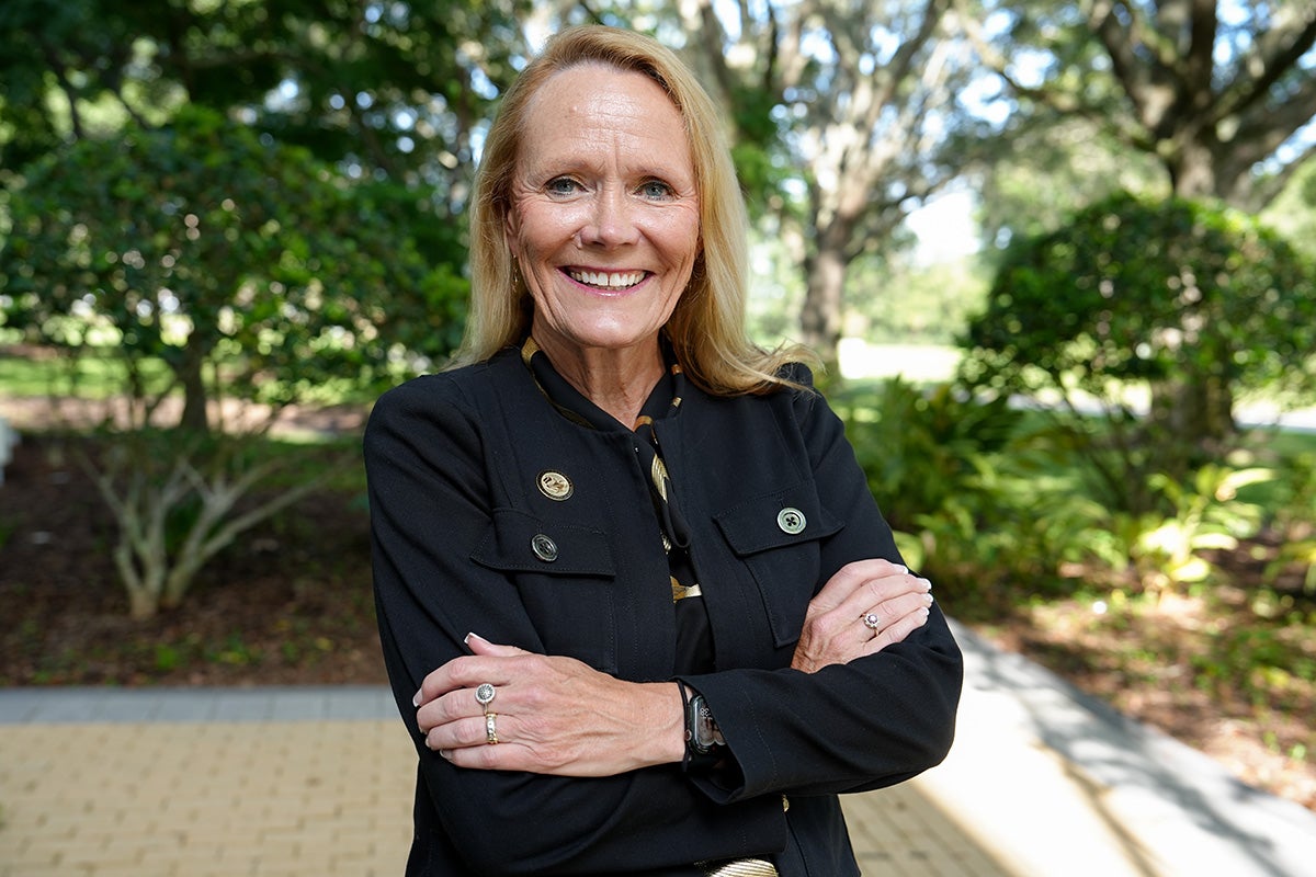 Blonde woman wearing black collared shirt poses with arms crossed outside with green trees behind her