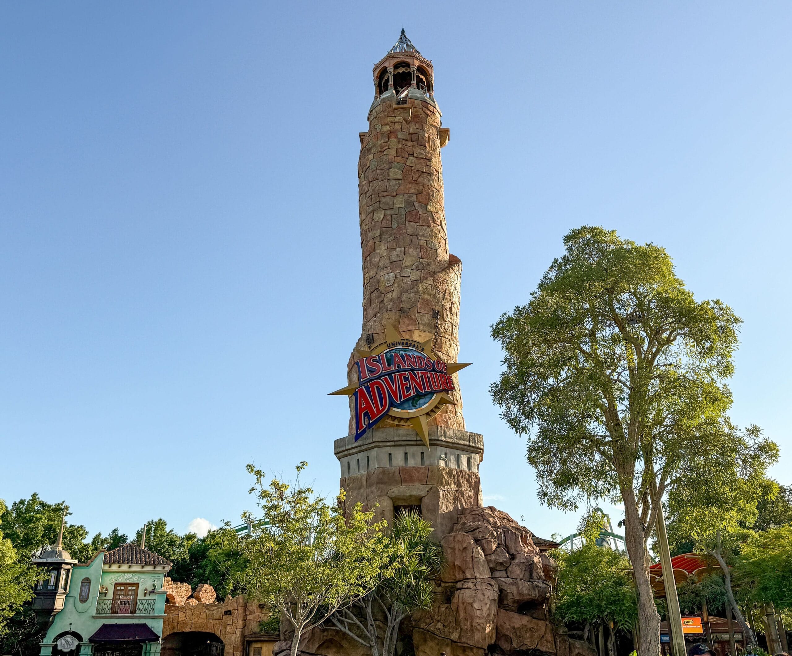 A tall stone lighthouse with a sign reading “Islands of Adventure” towers near trees and Camp Jurassic under a clear blue sky.
