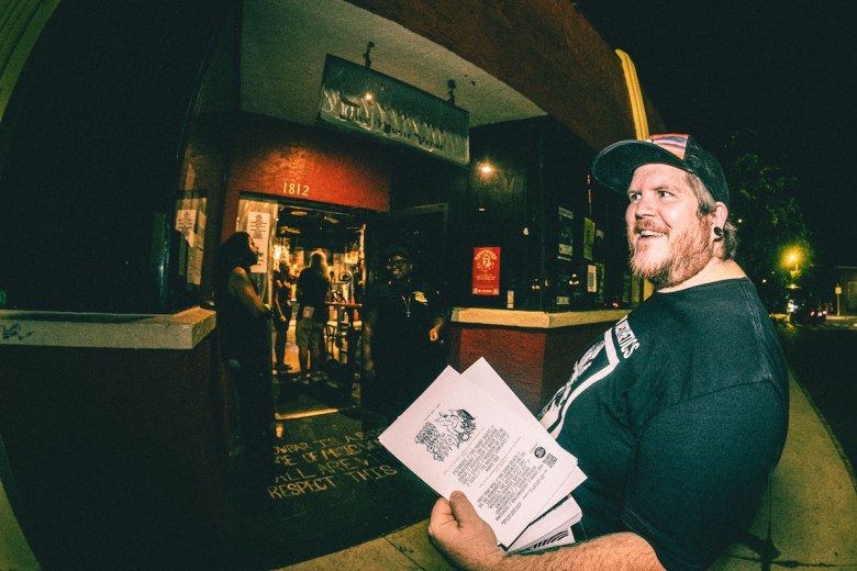 A wide-angle, low-light photo taken outside a music venue at night. A bearded man in a baseball cap and black t-shirt is smiling, looking off to the right while holding flyers. In the background, patrons are gathered near the red entrance door of the building, which has the address 1812 visible.