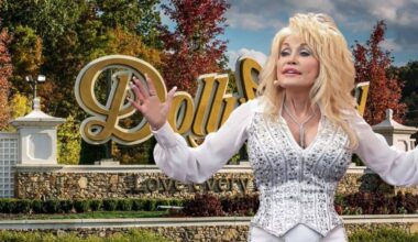 Dolly Parton in a sparkly white outfit stands with her arms outstretched in front of the Dollywood sign. The background features trees with autumn foliage and a stone wall below the sign that reads "Dollywood" with the tagline "Love every moment."
