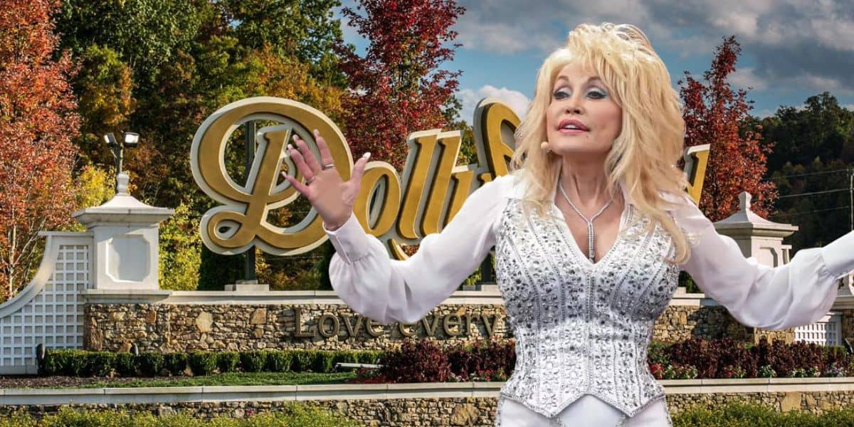 Dolly Parton in a sparkly white outfit stands with her arms outstretched in front of the Dollywood sign. The background features trees with autumn foliage and a stone wall below the sign that reads "Dollywood" with the tagline "Love every moment."