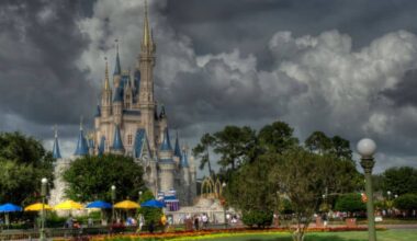 A large and ornate castle with spires, surrounded by lush greenery and a colorful garden with umbrellas, under dark, dramatic clouds in the sky. People are walking around the castle, enjoying the park.