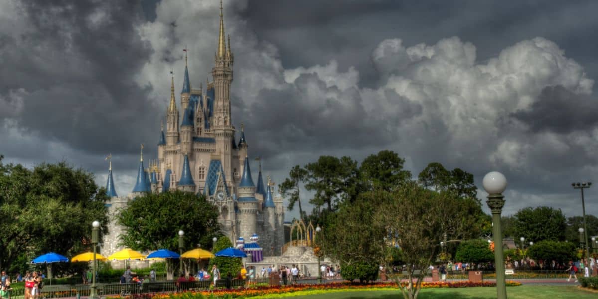 A large and ornate castle with spires, surrounded by lush greenery and a colorful garden with umbrellas, under dark, dramatic clouds in the sky. People are walking around the castle, enjoying the park.
