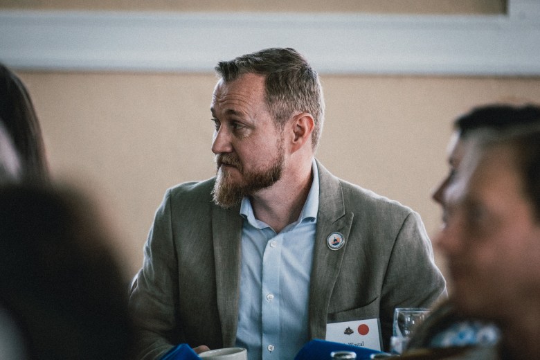 Hillsborough County Commissioner Josh Wostal sits at a table during a Tampa Tiger Bay Club event. He is viewed from the side, looking toward the speaker with a focused expression. He has a beard and short light-brown hair, wearing a tan blazer over a light blue button-down shirt. The background is softly blurred, showing other attendees in a professional banquet setting.