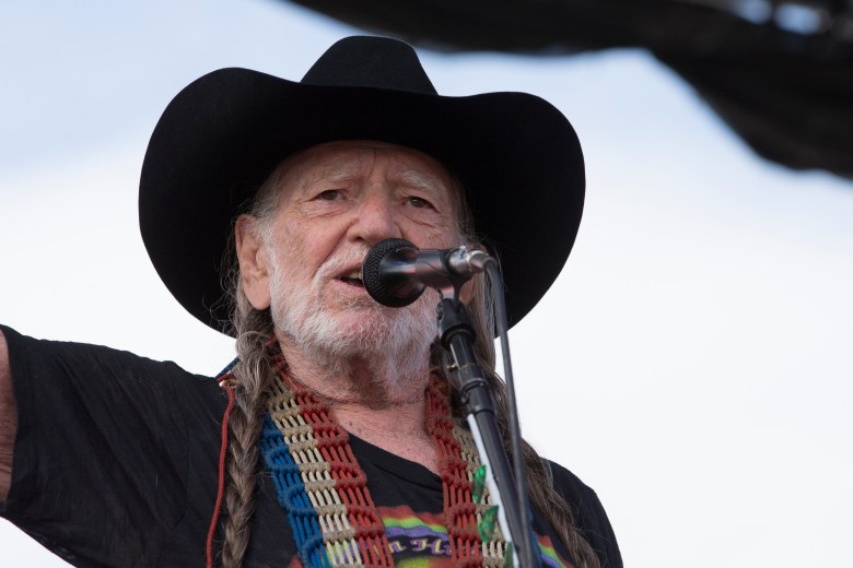 A low-angle close-up of country music legend Willie Nelson performing at a microphone. He is wearing his signature black cowboy hat and has his long grey hair in braids. A colorful, multi-strand beaded guitar strap is visible over his black t-shirt against a bright, slightly overexposed sky.