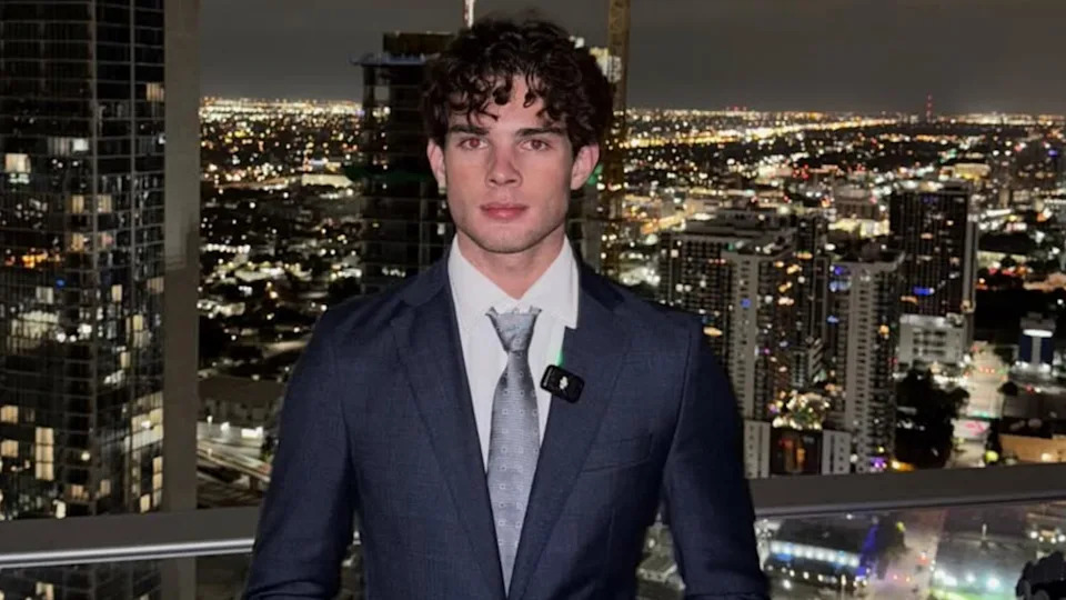 Clavicular in a suit stands on a balcony overlooking a cityscape at night, with buildings and lights in the background.