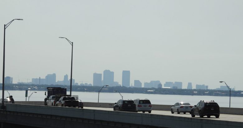 A wide-angle, slightly hazy daytime photograph looking across Old Tampa Bay toward the distant Downtown Tampa skyline. In the foreground, a multi-lane highway bridge is visible with several cars and a large truck traveling toward the city. Two tall, modern streetlights curve over the road from the left. The skyline is a soft grey silhouette against a pale, overcast sky, reflecting a typical humid afternoon in the Tampa Bay area.