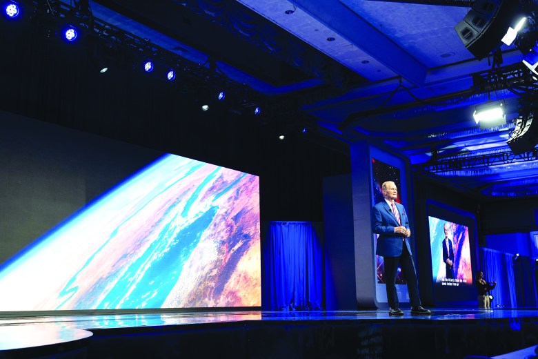 Bill Nelson giving a presentation on a brightly lit stage in front of a large screen displaying Earth from space.