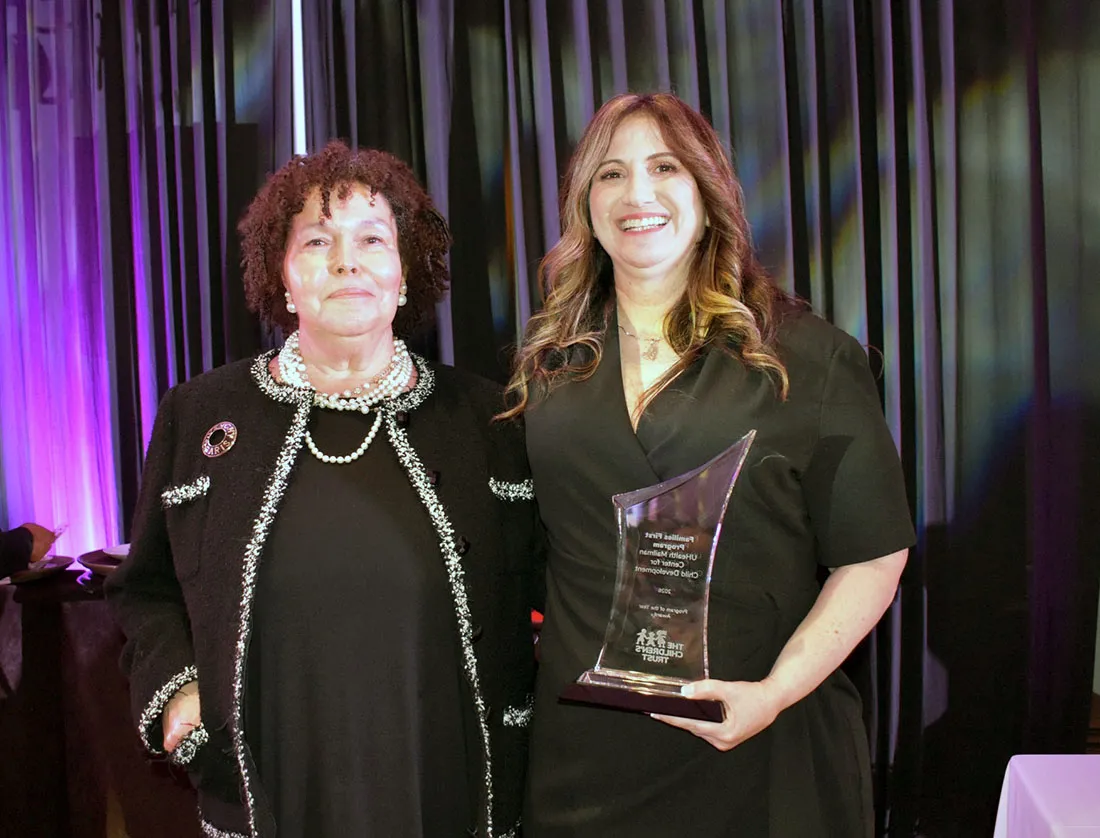 Dr. Elana Mansoor standing with an award presenter at an awards ceremony, with the person on the right holding a glass recognition award, photographed against a dark stage curtain with colored lighting.