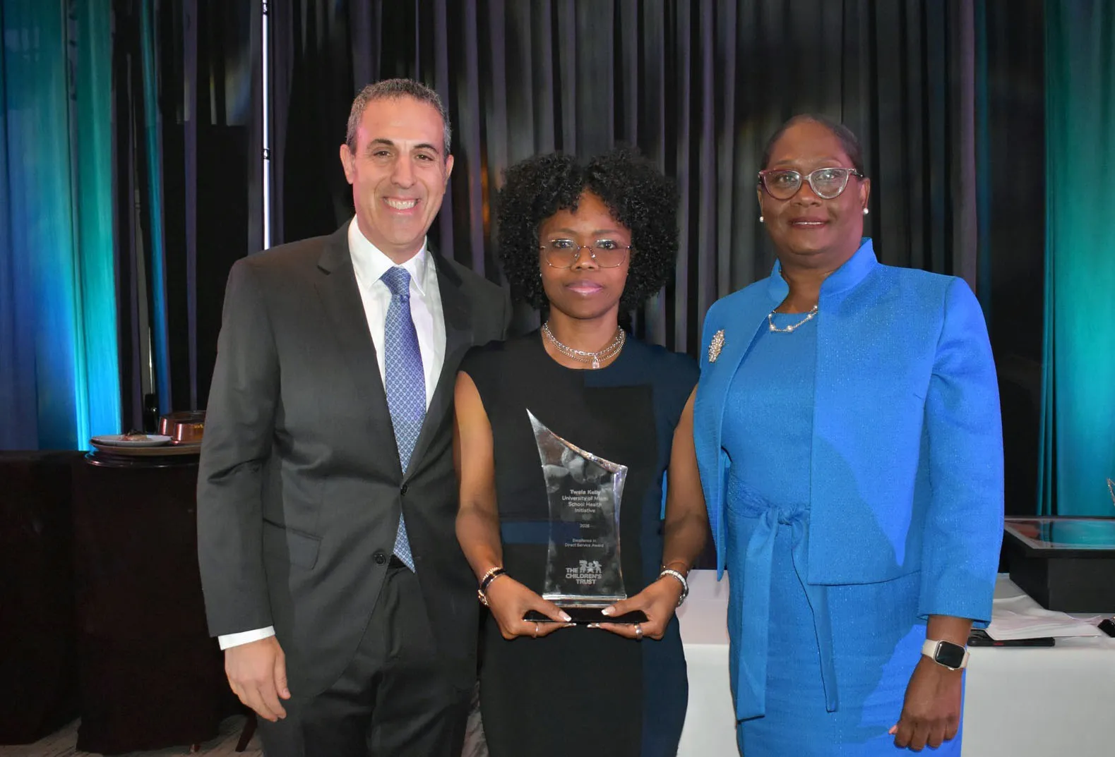 Twala Kelly (center) standing with two award presenters at an awards ceremony, with the person in the center holding a glass award, photographed in front of a dark stage backdrop with event lighting.