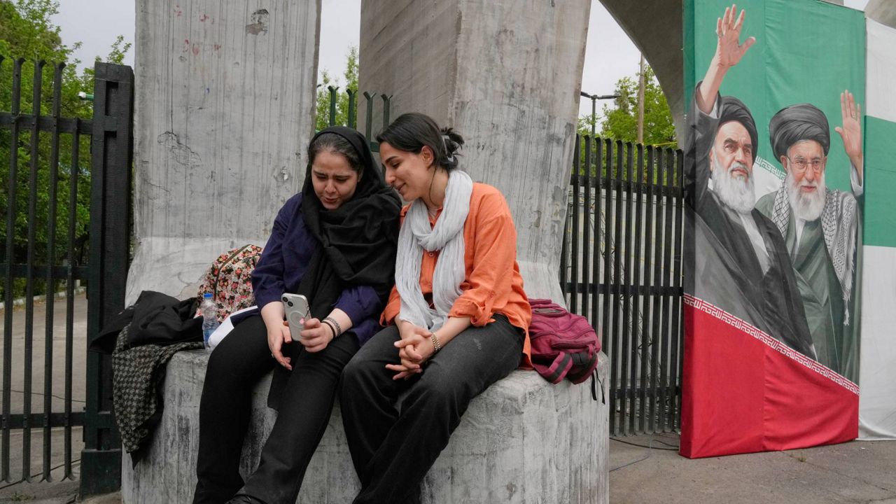 Women share a moment as they look at a smartphone at the main gate of the Tehran University as a banner shows portraits of the late Supreme Leader Ayatollah Ali Khamenei, right, and the late revolutionary founder Ayatollah Khomeini in Tehran, Iran, Sunday, April 19, 2026. (AP Photo/Vahid Salemi)