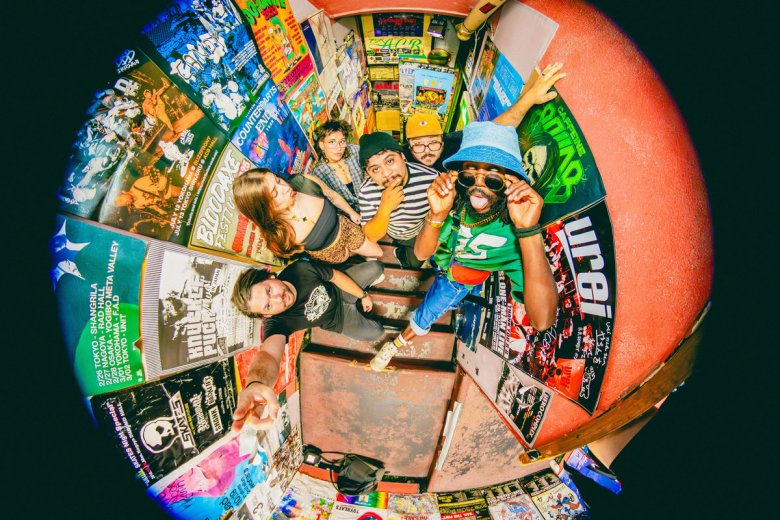 A high-angle fisheye lens photo of the band JER and their touring members standing in a cramped stairwell covered in colorful punk and ska gig posters. The members look up at the camera with various expressions, including Jeremy Hunter in a bright green shirt, blue bucket hat, and sunglasses, while the walls around them are a collage of concert flyers.