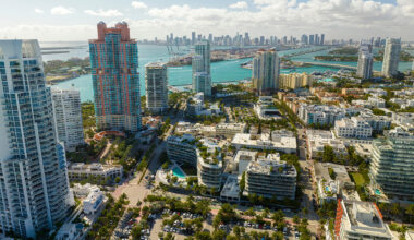 Aerial view of South Florida real estate with tall modern buildings, turquoise water, palm trees, and a distant skyline under a partly cloudy sky.