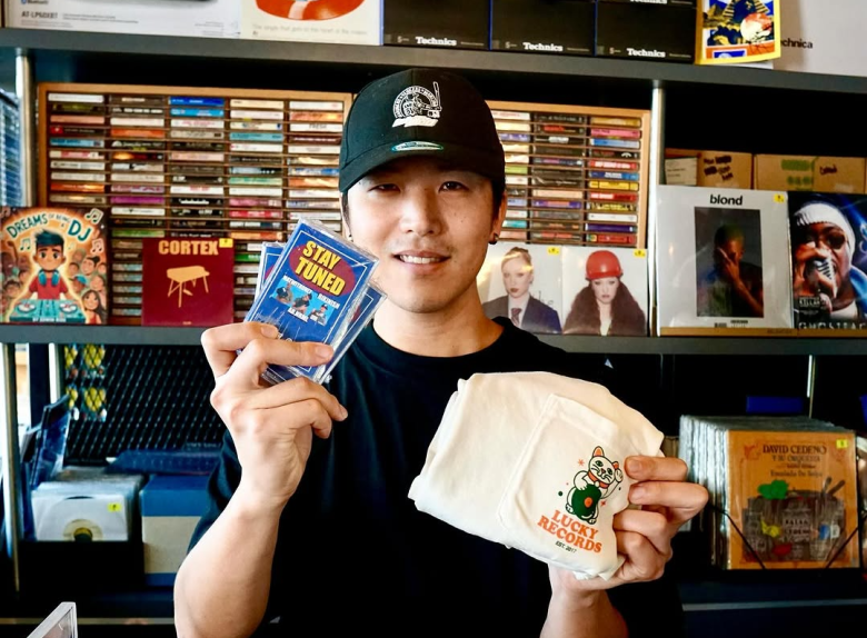 DJ and producer Rikinish smiling in a black baseball cap, holding a "Stay Tuned" cassette tape and a Lucky Records tote bag. He is standing in front of a shelf filled with vinyl records and CDs, including a copy of Frank Ocean’s "Blonde."