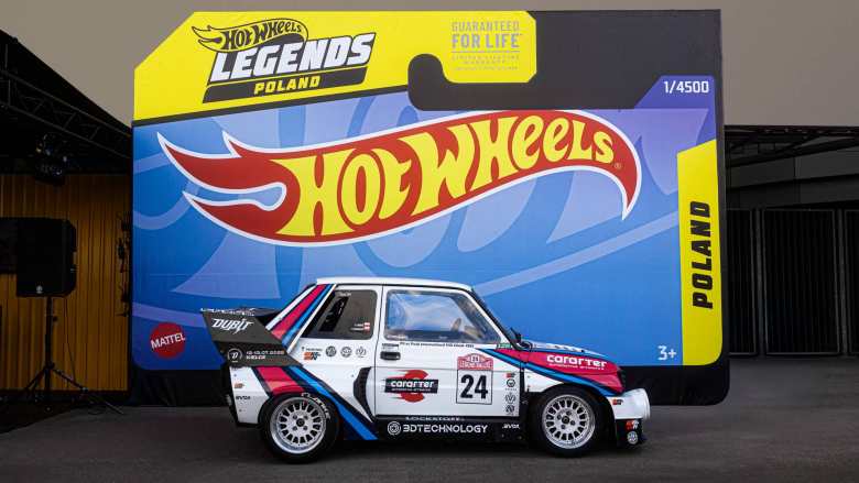 A custom-built, rally-themed white Fiat 126p (Maluch) parked in front of a giant Hot Wheels Legends Tour Poland display. The car features a wide-body kit, a massive rear wing, and a red, blue, and black 'Pikes Peak' style livery. It is adorned with various sponsor decals including 'cararter,' '3DTECHNOLOGY,' and 'Dubiit.' The backdrop is designed to look like a Hot Wheels toy car package, complete with the iconic flame logo and the text 'Guaranteed for Life.'