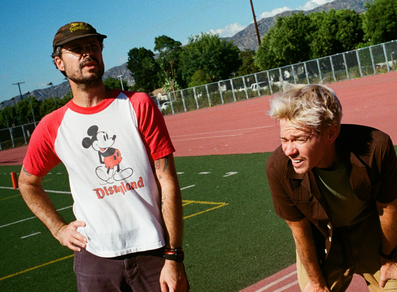 Nate Ruess and Sam Means of the band The Format standing on an outdoor athletic track under a bright blue sky. On the left, Nate wears a brown baseball cap, glasses, and a red-and-white Mickey Mouse "Disneyland" raglan shirt. On the right, Sam, with bleached blonde hair, leans forward in a brown button-down shirt. The background features green trees and distant mountains.