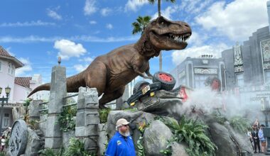 A lifelike T. rex towers over a crushed car at Universal Studios Florida, with smoke rising and a staff member nearby.