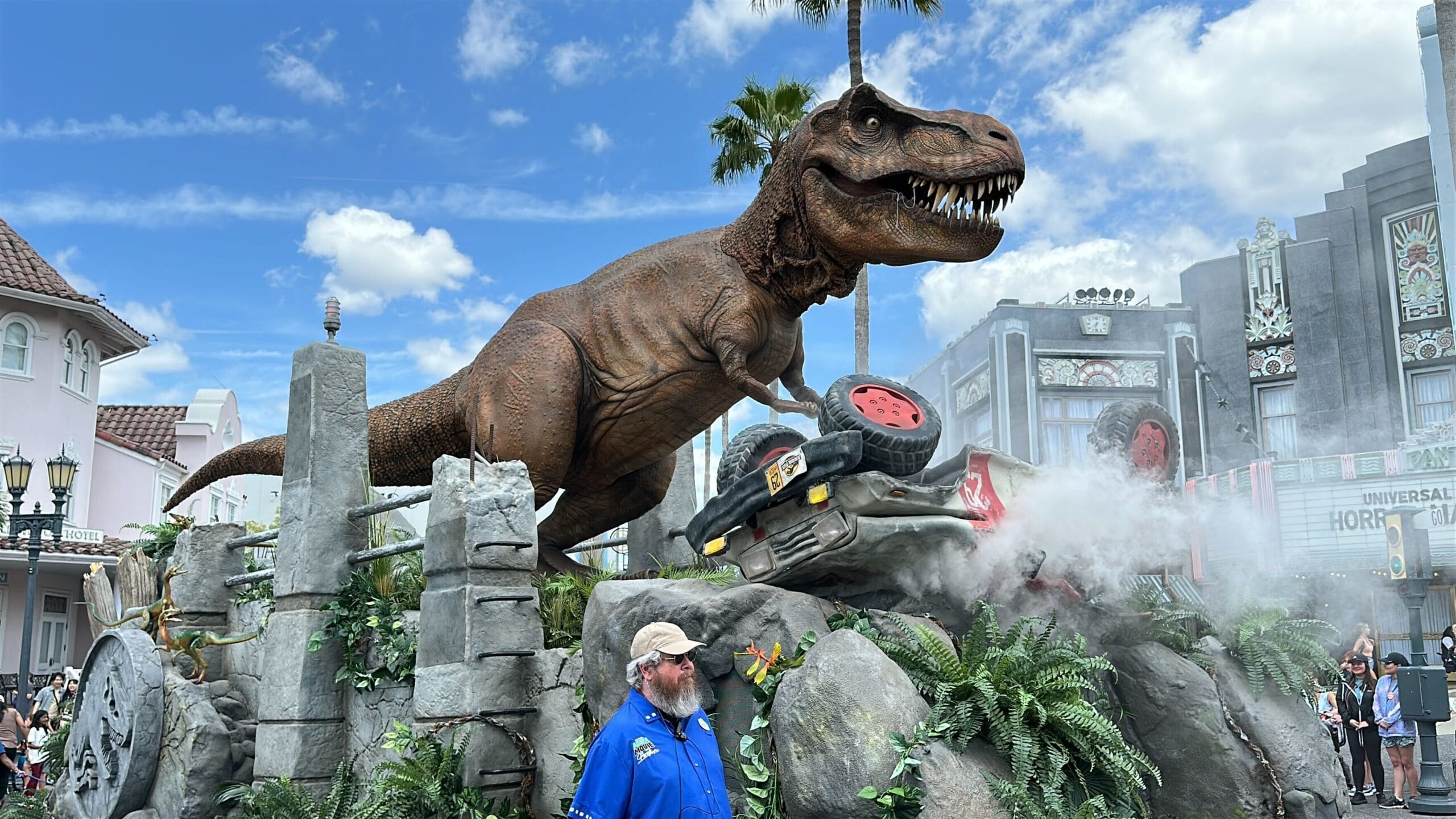 A lifelike T. rex towers over a crushed car at Universal Studios Florida, with smoke rising and a staff member nearby.
