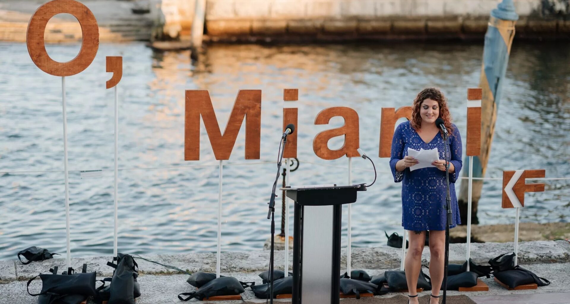 a person reads a poem in front of the water and a backdrop that reads, "O, Miami"