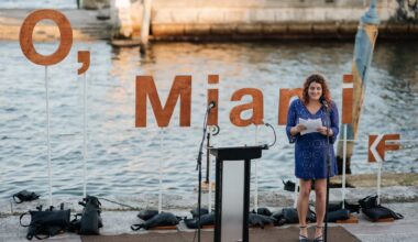 a person reads a poem in front of the water and a backdrop that reads, "O, Miami"