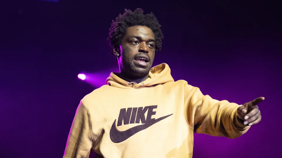 Kodak Black in a Nike hoodie performs on stage with purple lighting in the background, pointing forward.