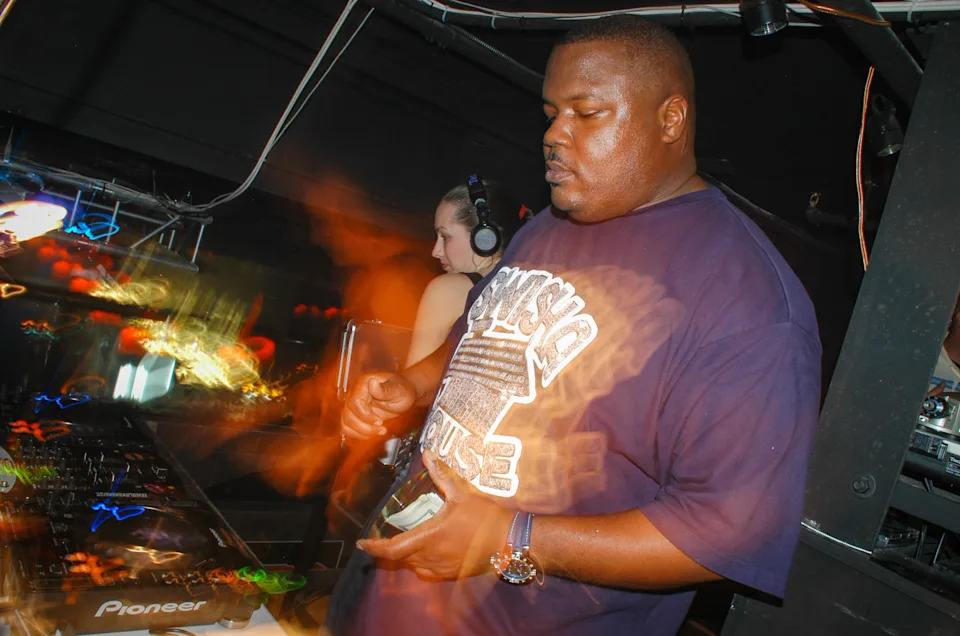 Michael "5000" Watts performing in a dimly lit club, he is wearing headphones, surrounded by colorful lights. Michael "5000" Watts died in 2026.