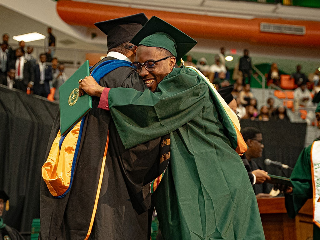 A student at graduation. A male graduate hugging an administrator.