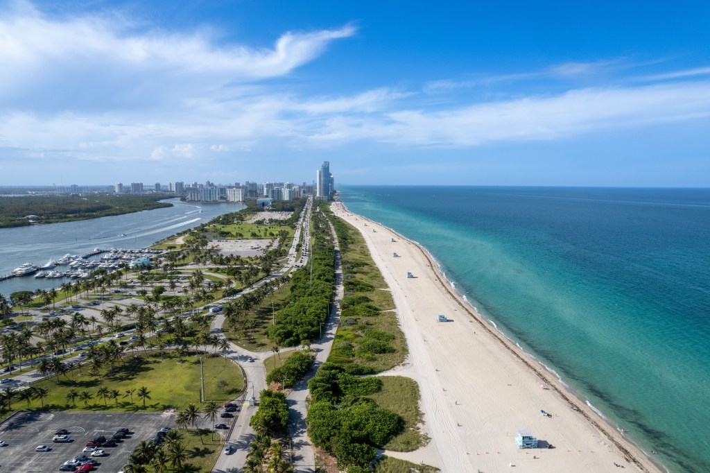 Aerial view of Haulover Beach in Miami with a long sandy beach, turquoise ocean, a road lined with palm trees, and a marina.