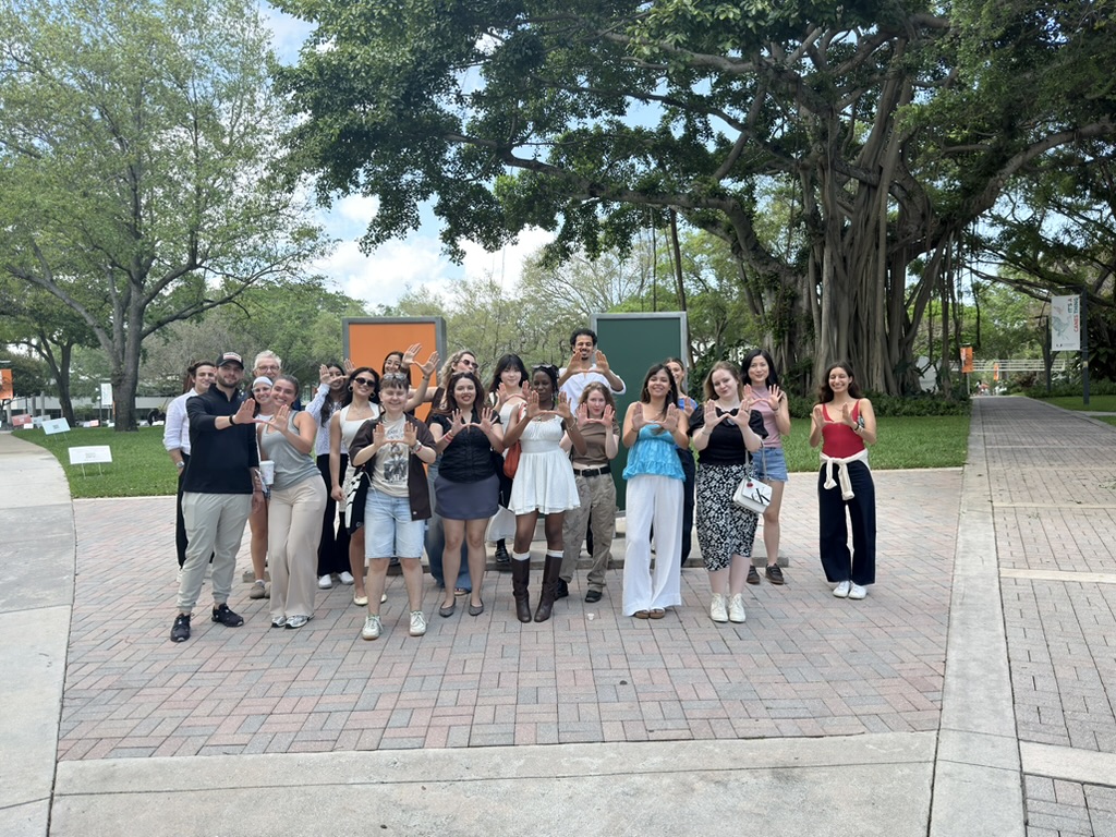 A diverse group of individuals stands together outdoors, smiling and posing for the photo. They are in a park-like setting with trees and greenery in the background. Many participants are making hand gestures, and some are dressed casually, reflecting a relaxed atmosphere. The ground is paved with bricks, and there are informational signs visible in the background.