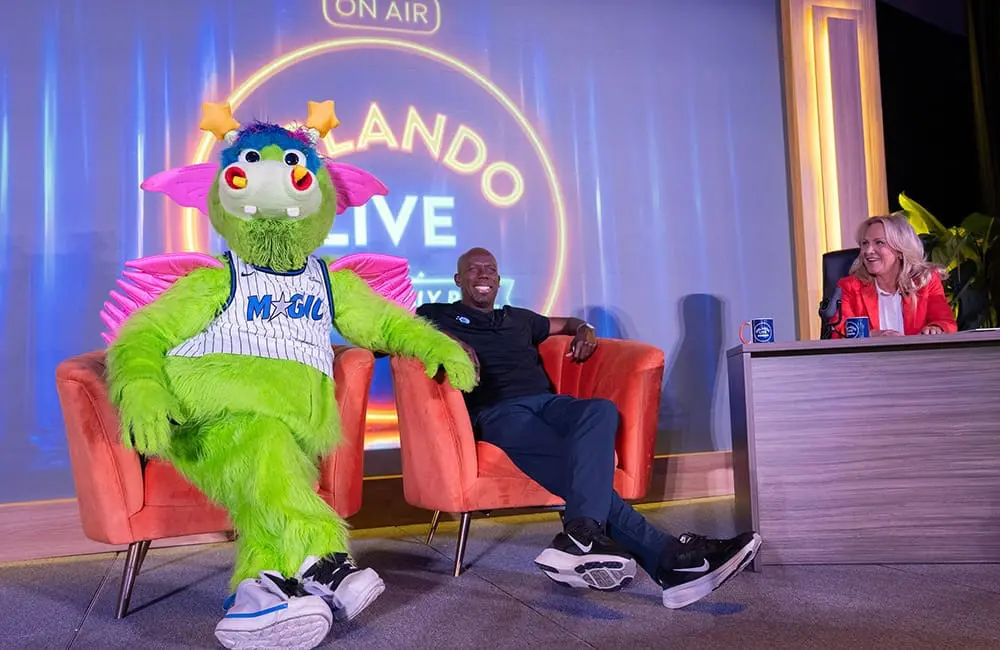 Orlando Magic's Stuff the Magic Dragon, retired basketball player Bo Outlaw and Visit Orlando President & CEO Casandra Matej at the Visit Orlando press conference at IPW 2026.