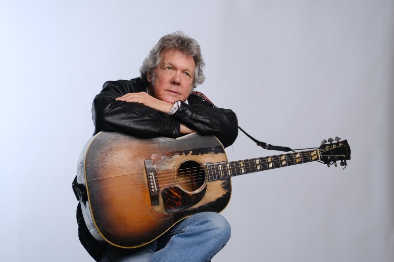 A close-up studio portrait of the singer-songwriter Steve Forbert against a plain, light-colored background. They are wearing a classic black leather jacket, with one hand resting gently across their chest. Wisps of white hair are partially visible against the bright backdrop. The lighting is soft and even, highlighting the texture of the leather and their relaxed, contemplative posture.