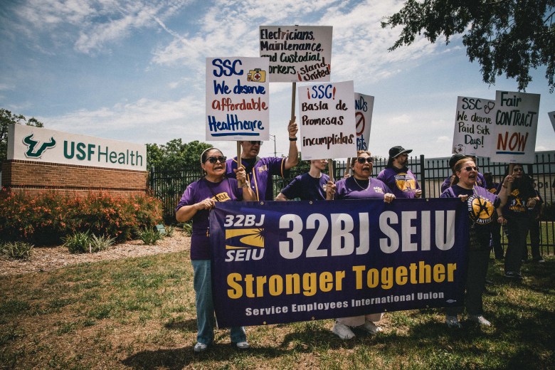 A daytime photograph of a group of workers holding a labor rally outside the USF Health campus in Tampa. In the foreground, several individuals wearing matching purple t-shirts are holding a large purple banner that reads '32BJ SEIU Stronger Together.' They are raising various handmade and printed protest signs with messages including 'SSC We deserve affordable Healthcare,' 'Electricians, Maintenance and Custodial workers stand United,' and the Spanish phrase 'SSC! Basta de Promesas incumplidas.' Behind the demonstrators, a brick monument sign displaying the 'USF Health' logo is visible against a bright blue sky with scattered clouds.