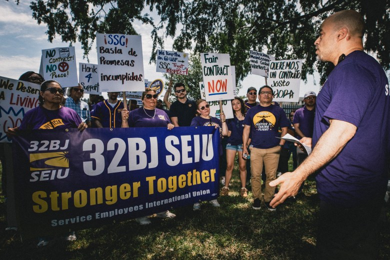 A daytime photograph of Onex Cortez standing in profile in the foreground during a labor rally outside the University of South Florida. They are wearing a dark purple t-shirt and gesturing with one hand toward a crowd of demonstrators. In the background, the group is gathered on a grassy, tree-shaded area wearing matching purple shirts. They are holding a large banner that reads '32BJ SEIU Stronger Together' alongside the Service Employees International Union logo, and are raising protest signs with messages including 'Fair Contract NOW' and the Spanish phrase 'SSC! Basta de Promesas incumplidas.'