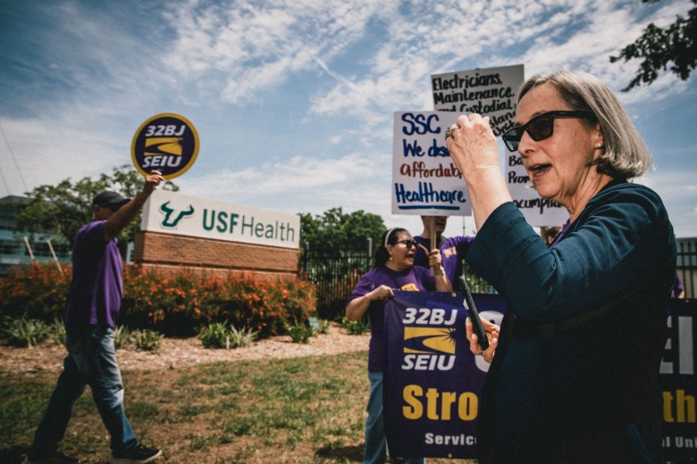 A daytime photograph of Helene O'Brien speaking into a handheld microphone at a labor rally outside the USF Health campus in Tampa. O'Brien, wearing sunglasses and a dark long-sleeve top, stands in the foreground in profile. Behind them, individuals wearing matching purple t-shirts are holding a large banner that reads '32BJ SEIU Stronger Together.' Demonstrators are raising protest signs with messages including 'Electricians, Maintenance, Custodial...' and 'SSC We deserve Affordable Healthcare.' A brick monument sign displaying the 'USF Health' logo is visible in the background under a bright sky with scattered clouds.
