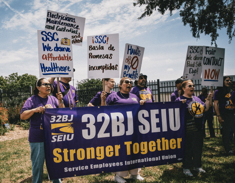 A daytime photograph of a group of workers holding a labor rally outside the USF Health campus in Tampa. In the foreground, several individuals wearing matching purple t-shirts are holding a large purple banner that reads '32BJ SEIU Stronger Together.' They are raising various handmade and printed protest signs with messages including 'SSC We deserve affordable Healthcare,' 'Electricians, Maintenance and Custodial workers stand United,' and the Spanish phrase 'SSC! Basta de Promesas incumplidas.'