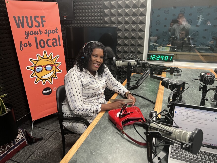 Black woman in white pattern blouse and shoulder length hair with headphones on, sits at a table while talking into a large microphone in a radio studio. Behind her is a tall orange sign with the words USF Your Spots for Local.