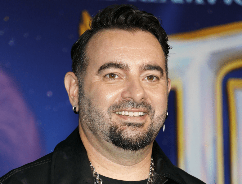 A close-up portrait photograph of musician Chris Kirkpatrick smiling warmly at the camera. They have short dark hair, a neatly trimmed beard with touches of gray, and are wearing a black jacket layered over a black top, accented by a thick silver chain necklace. The background is softly blurred with a shallow depth of field, revealing abstract shapes in dark blue and bright yellow, indicative of a brightly lit event or premiere setting.