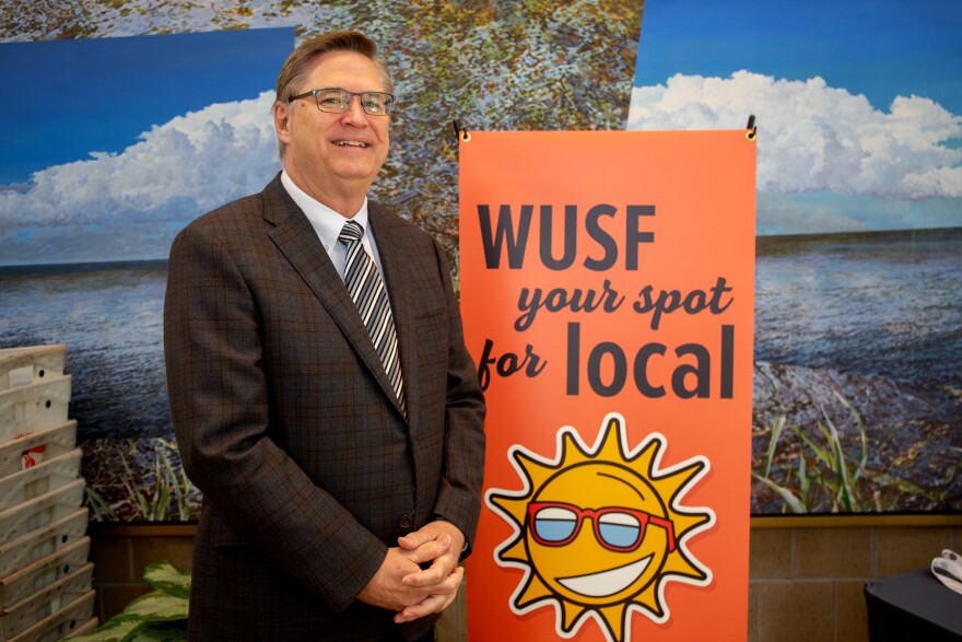 Man in suit and tie wearing glasses stands in front of WUSF Your Spot for Local sign.