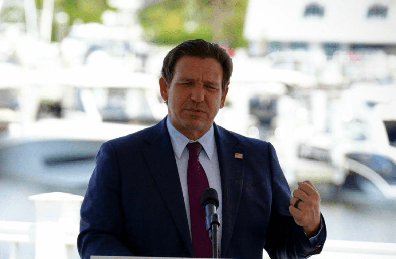 A bright, daytime outdoor photograph of Florida Governor Ron DeSantis speaking at a press conference. Standing behind a podium and a white railing, they are wearing a dark blue suit, a light blue shirt, and a dark red tie. They are gesturing with their raised left hand while speaking. The background features a softly blurred marina with several white boats docked on the water.