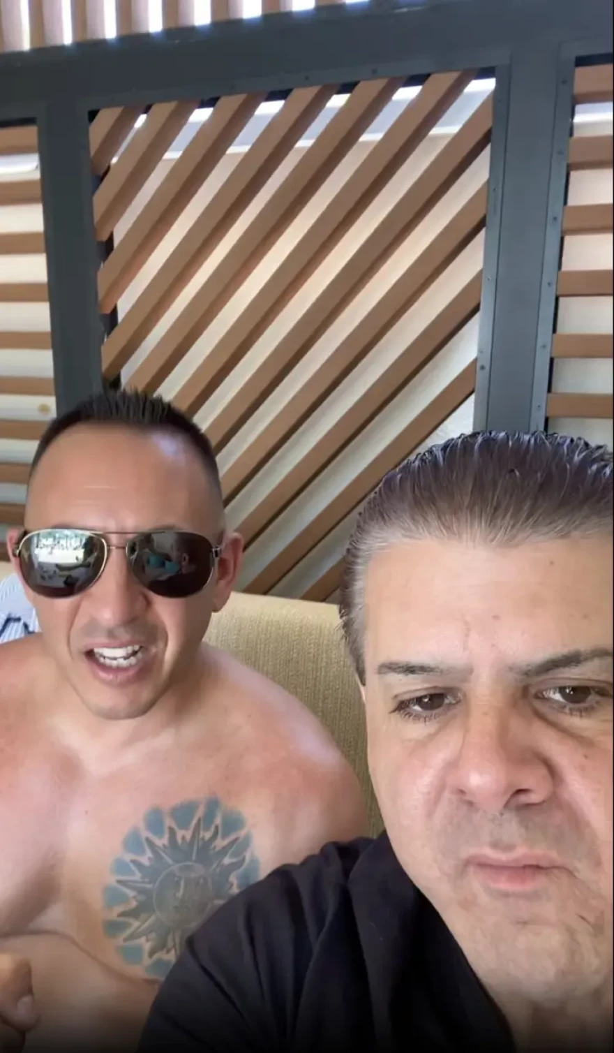 One shirtless man with tattoo on chest sits next to smirking man with slicked back hair.