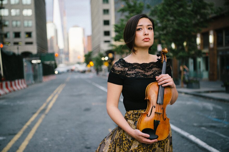 A young woman in a dark lace blouse with a printed skirt holds a violin while standing on the pavement.