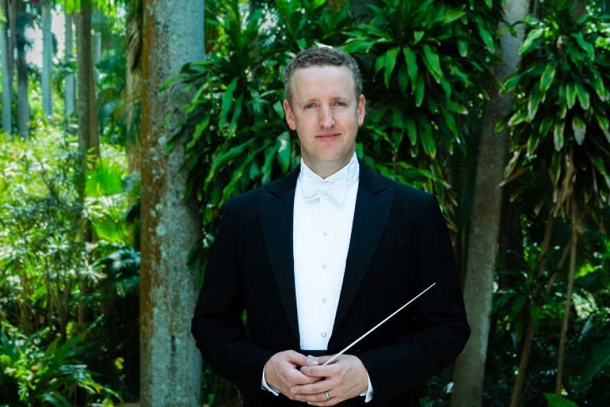 A man in a tuxedo stands in the midst of green trees.