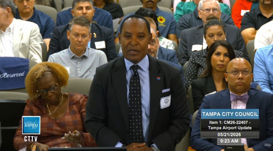Black man in a blue suit looking straight ahead and talking, with people in a meeting room behind him. Photo is a video screengrab