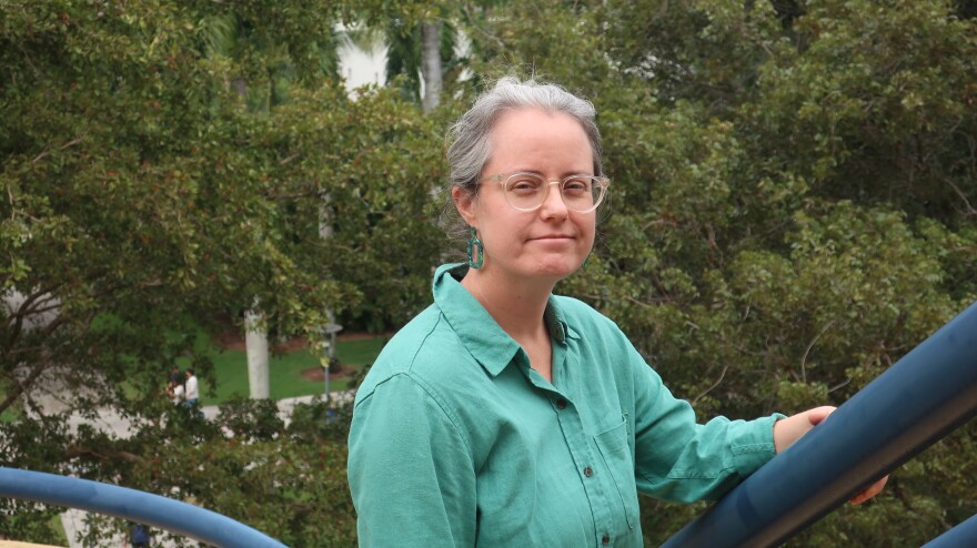 Katie Rainwater, a woman in a green shirt and green earing, translucent eyeglasses and grey hair, looks at the camera from a staircase. Greenery is below her.