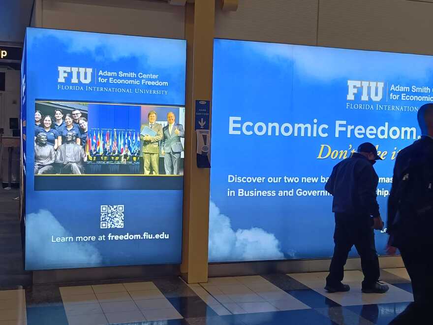 A man walks past a digital billboard at Reagan International Airport advertising the Adam Smith Center for Economic Freedom at FIU. On the screen is an image of Argentinian President Javier Milei embracing Carlos Diaz-Rosillo.
