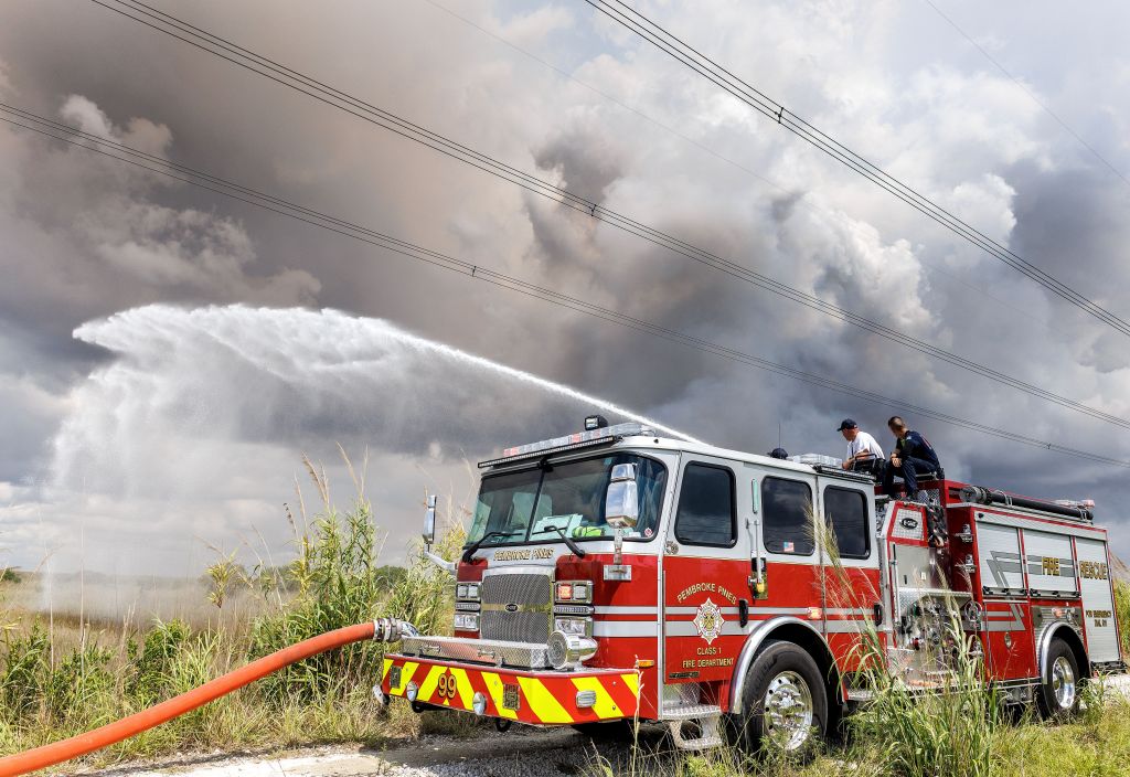 Firefighters spraying water from a fire truck with smoke in the background.