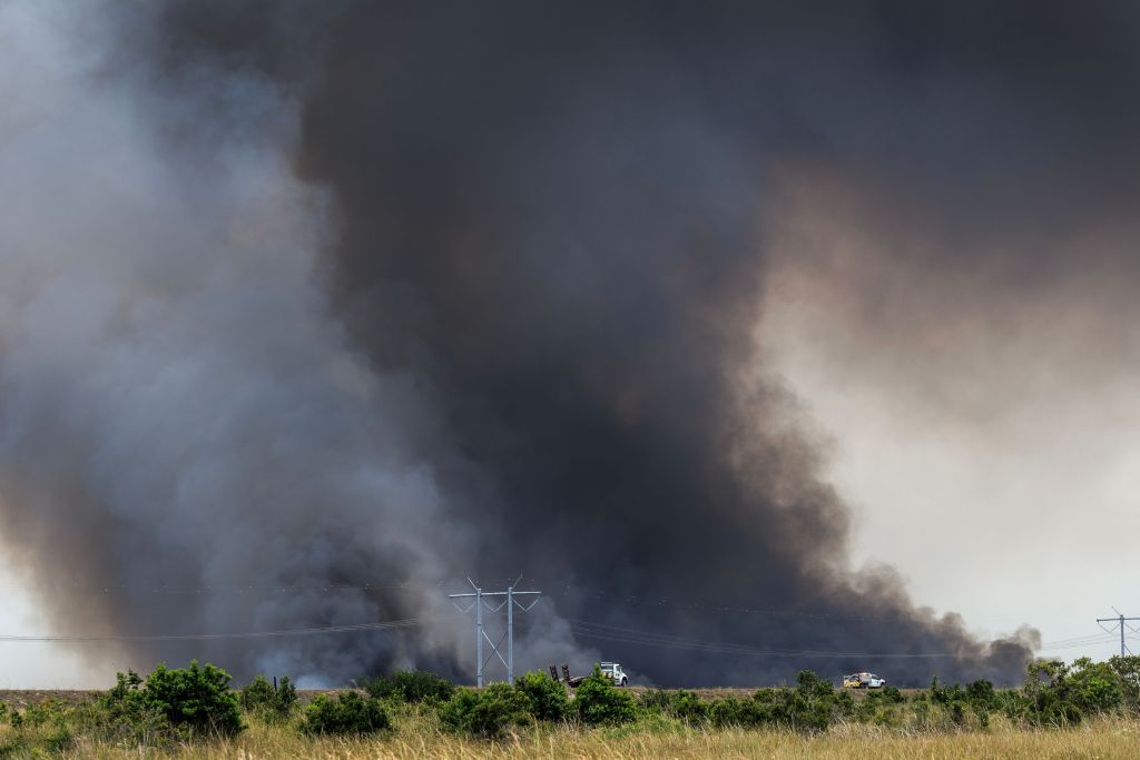 Thick black and gray smoke rises from a wildfire in Pembroke Pines, Florida, with power lines and two emergency vehicles visible in the distance.