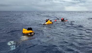 Two people in wet suits assist two other people with life preservers swimming in ocean into a recovery basket as a rescue helicopter hovers above.