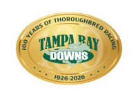 The 100 years anniversary logo for Tampa Bay Downs. The logo is a golden, oval emblem, with thin outlines surrounding the circumference of the emblem that make the vector look like shining gold. Dark green curving text along the outside of the logo reads, “100 Years of Thoroughbred Racing, 1926 - 2026.” In the middle of the logo, Tampa Bay Downs standard brand logo (consisting of a galloping racehorse and jockey, overlapping the name “Tampa Bay Downs” in dark and light green) is centered overtop a large “100,” which is partially transparent and fades into the gold of the logo.