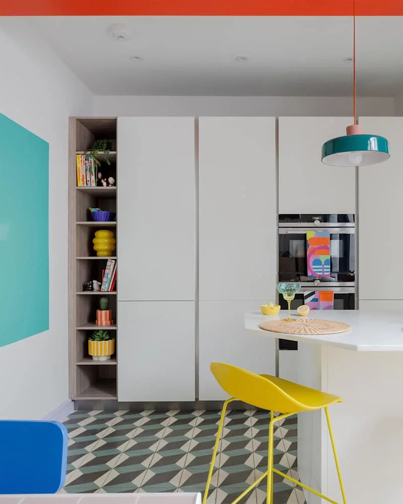 Bright kitchen featuring white cabinets, colorful decor, geometric floor tiles, and a yellow bar stool.